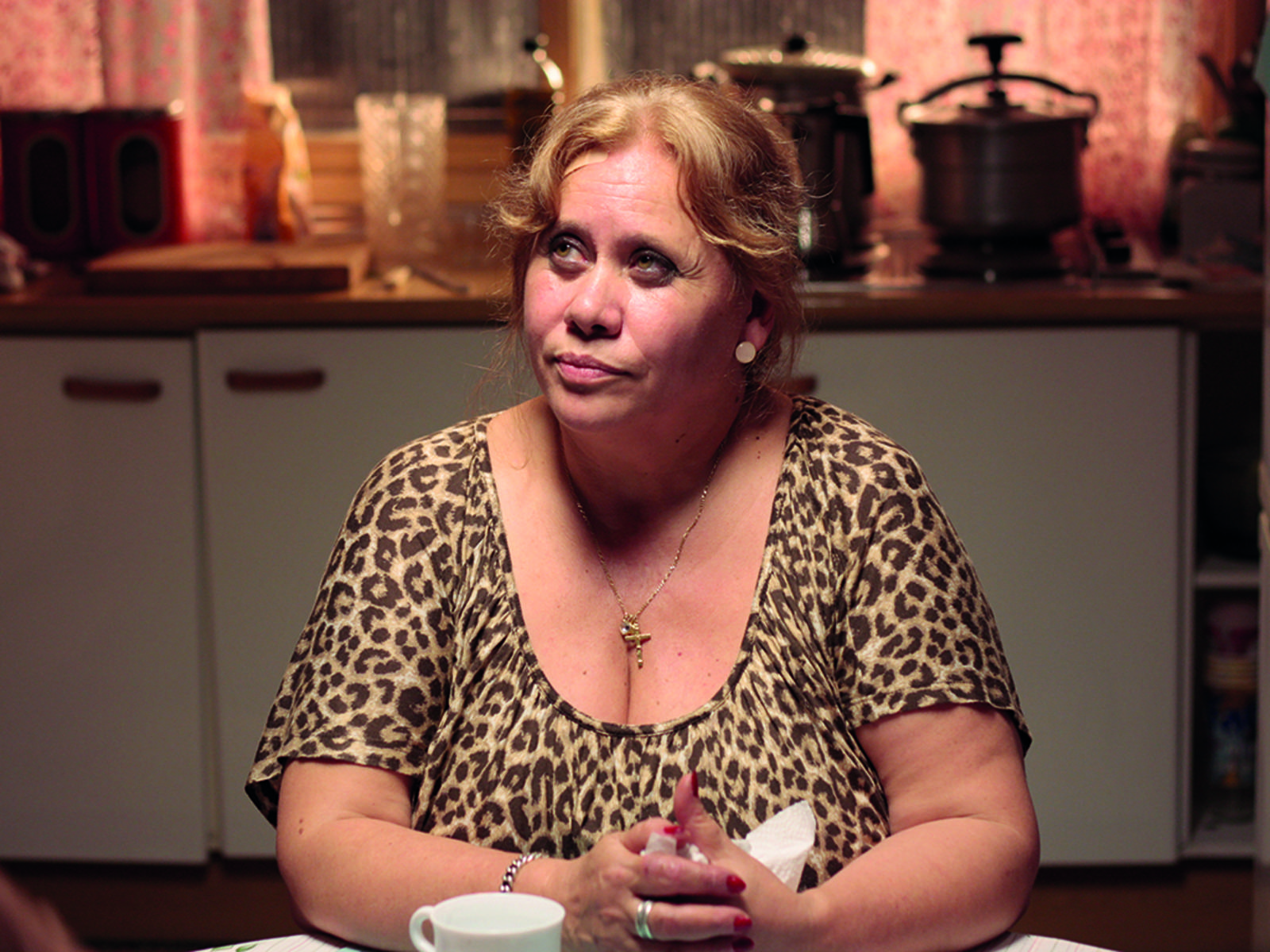Eine fast 60jährige Frau sitzt an einem Küchentisch. Sie ist blond und trägt ein Shirt in Leopardenfelloptik. Im Hintergrund eine Küchenzeile.